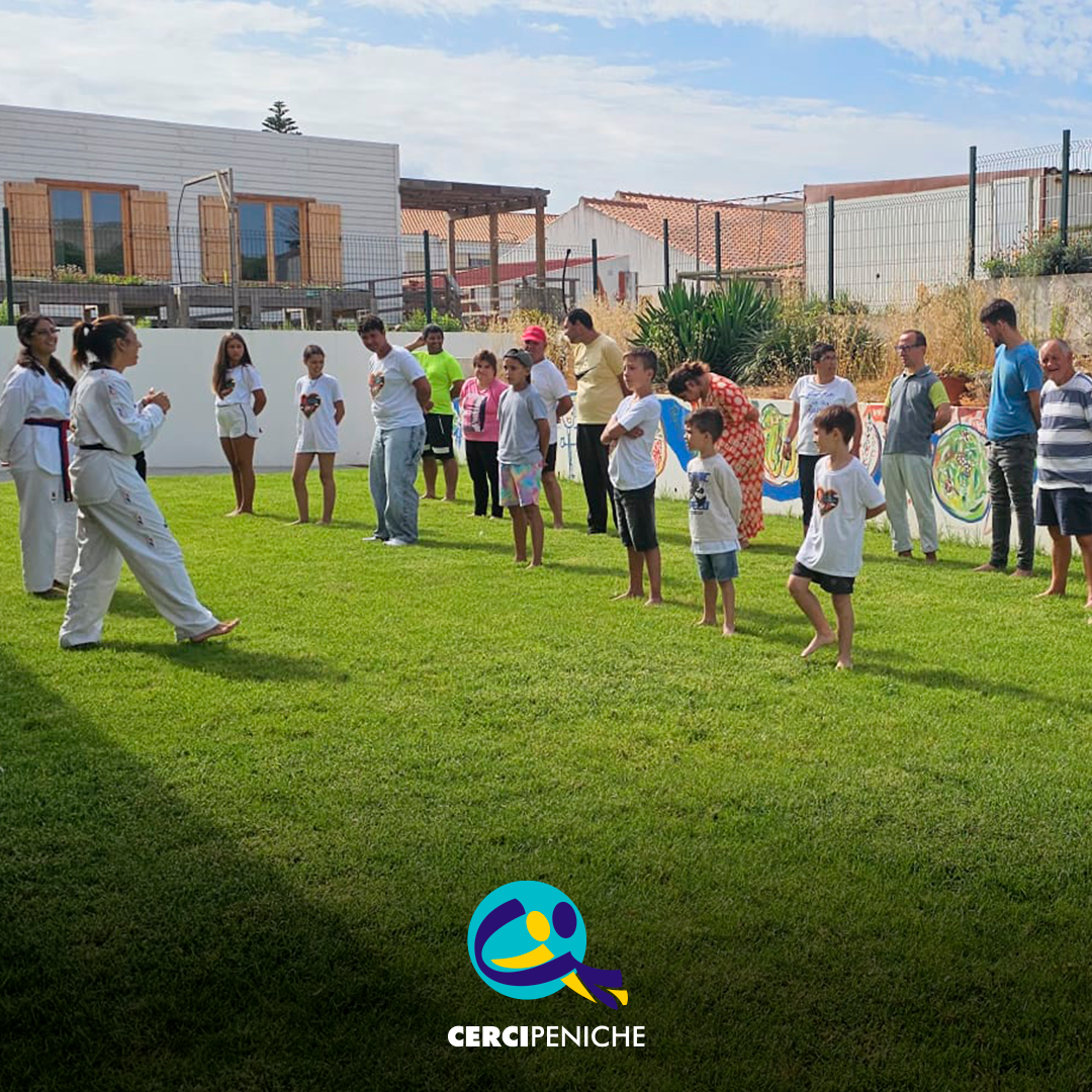Num espaço exterior relvado da Cercipeniche, um grupo de pessoas apoiadas participa numa atividade de Taekwondo, acompanhando os movimentos demonstrados por duas instrutoras. Logo da Cercipeniche.