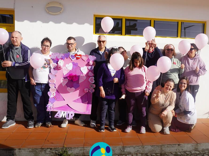 Grupo de pessoas apoiadas posa para uma fotografia no exterior do Cerisc. Várias pessoas seguram balões cor-de-rosa, enquanto outras apresentam o laço decorado com corações, flores, mensagens de sensibilização e um grande laço cor-de-rosa. Na parte inferior da imagem encontra-se o logótipo da Cercipeniche.
