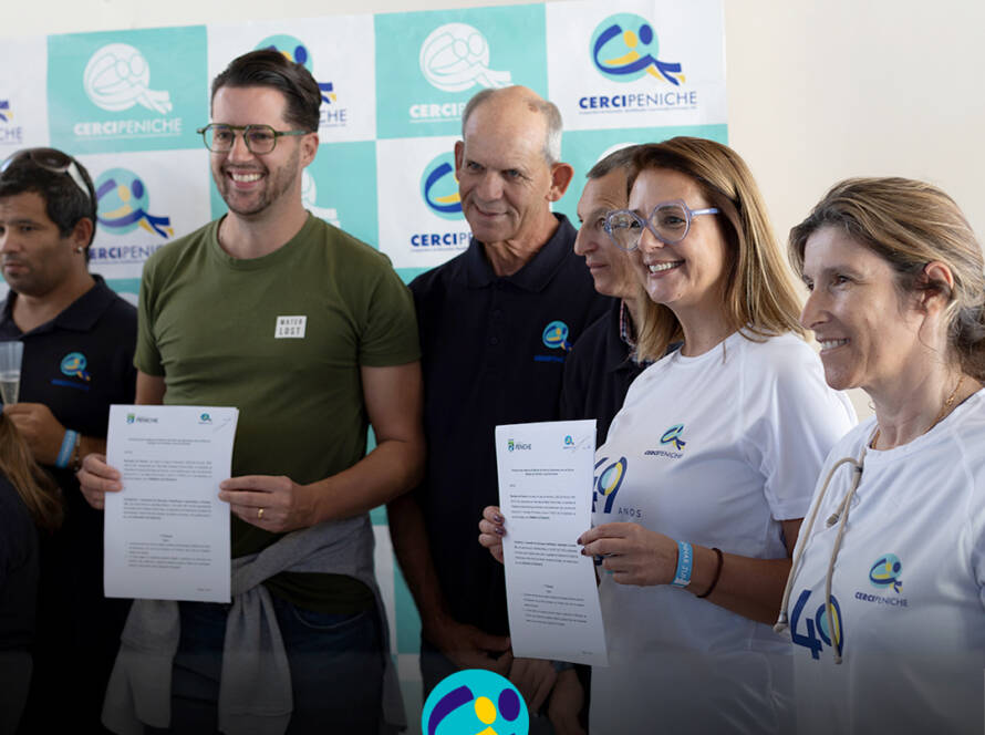 Grupo de seis pessoas posa para a fotografia em frente a um painel com os logótipos da Cercipeniche. Um homem de t-shirt verde e uma mulher com a t-shirt comemorativa dos 49 anos seguram documentos, enquanto os restantes participantes sorriem. Na parte inferior da imagem encontra-se o logótipo da Cercipeniche.