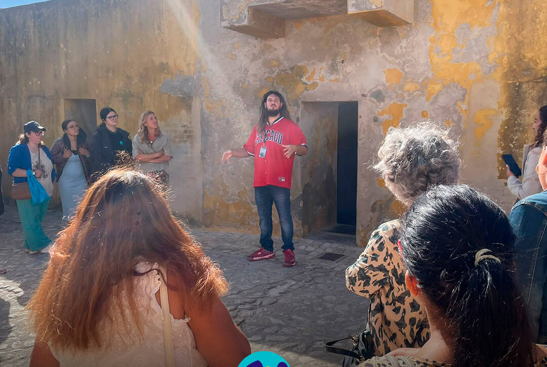 Dylan Almeida explica aos participantes diferentes aspetos do espaço e da sua história durante a visita guiada no exterior do Museu Nacional da Resistência e Liberdade. Na parte inferior da imagem encontra-se o logótipo da Cercipeniche.