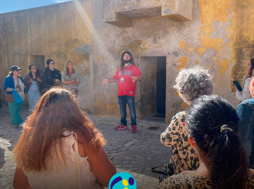Dylan Almeida explica aos participantes diferentes aspetos do espaço e da sua história durante a visita guiada no exterior do Museu Nacional da Resistência e Liberdade. Na parte inferior da imagem encontra-se o logótipo da Cercipeniche.