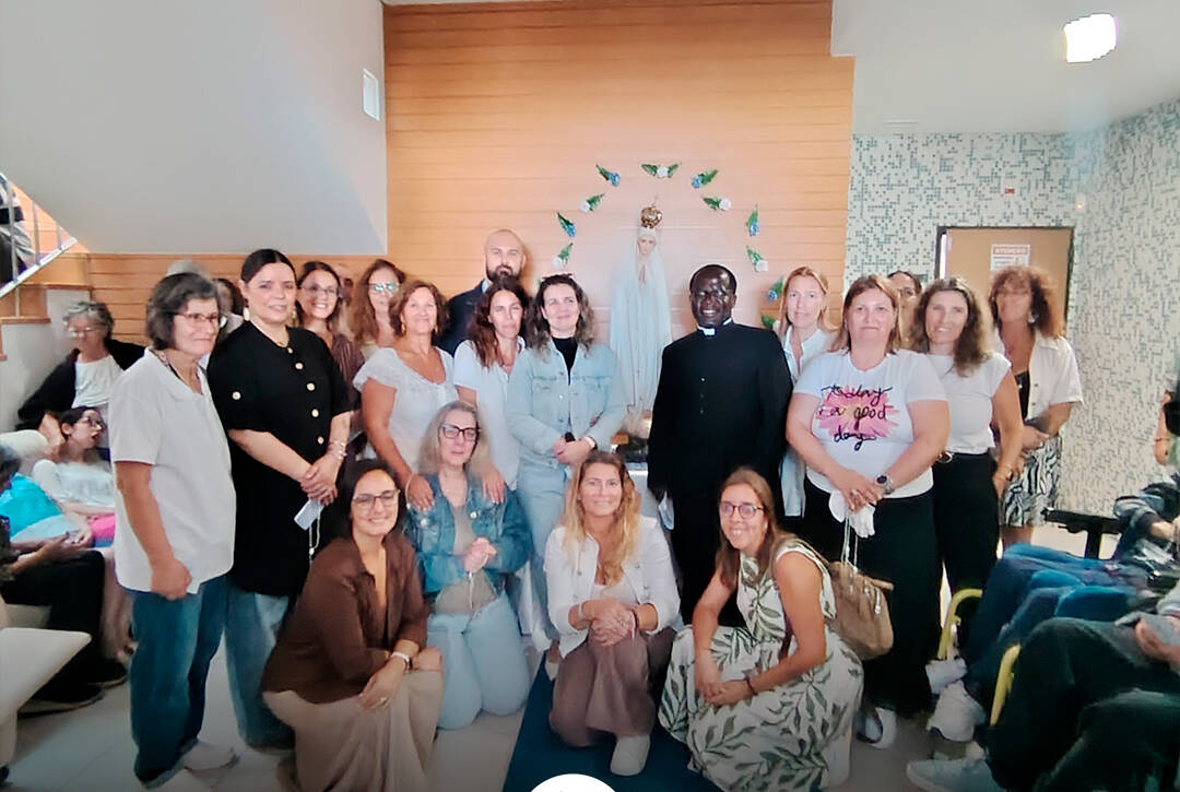 Um grupo de pessoas posa para uma fotografia no interior do Lar Porto de Abrigo, durante a visita de Nossa Senhora de Fátima Peregrina. Algumas pessoas estão de pé e outras agachadas. Ao centro encontra-se um sacerdote, vestido de preto, e ao fundo vê-se a imagem de Nossa Senhora. Na parte inferior da imagem encontra-se o logótipo da Cercipeniche.