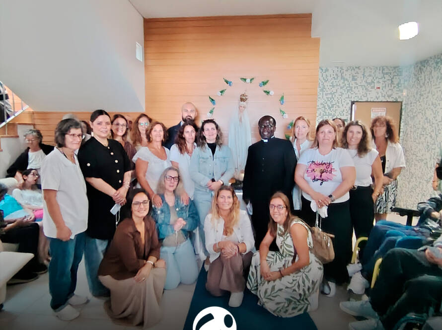 Um grupo de pessoas posa para uma fotografia no interior do Lar Porto de Abrigo, durante a visita de Nossa Senhora de Fátima Peregrina. Algumas pessoas estão de pé e outras agachadas. Ao centro encontra-se um sacerdote, vestido de preto, e ao fundo vê-se a imagem de Nossa Senhora. Na parte inferior da imagem encontra-se o logótipo da Cercipeniche.