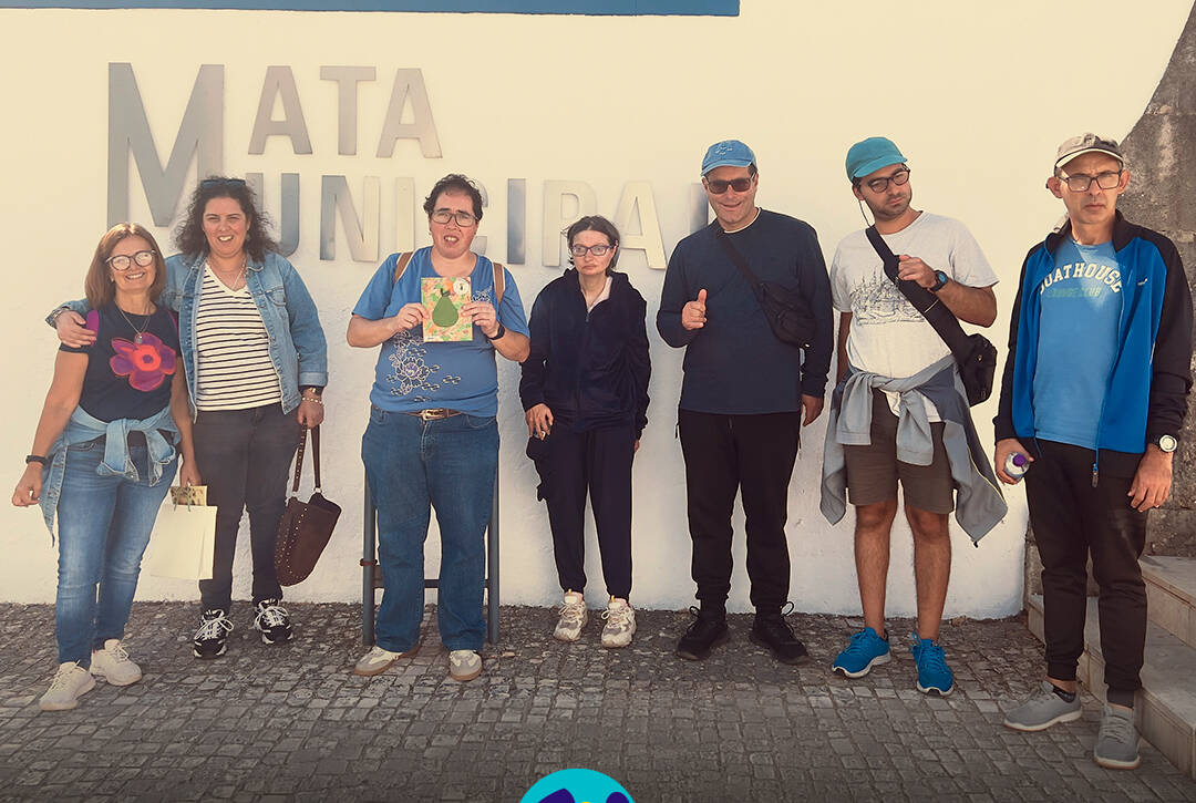 6 pessoas apoiadas e monitor da Cercipeniche posam para uma fotografia de grupo junto à entrada da Mata Municipal do Bombarral. Ao fundo, vê-se uma parede branca com a inscrição «Mata Municipal». Na parte inferior da imagem encontra-se o logótipo da Cercipeniche.