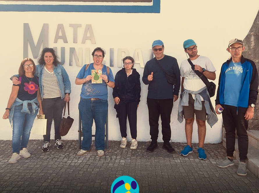 6 pessoas apoiadas e monitor da Cercipeniche posam para uma fotografia de grupo junto à entrada da Mata Municipal do Bombarral. Ao fundo, vê-se uma parede branca com a inscrição «Mata Municipal». Na parte inferior da imagem encontra-se o logótipo da Cercipeniche.