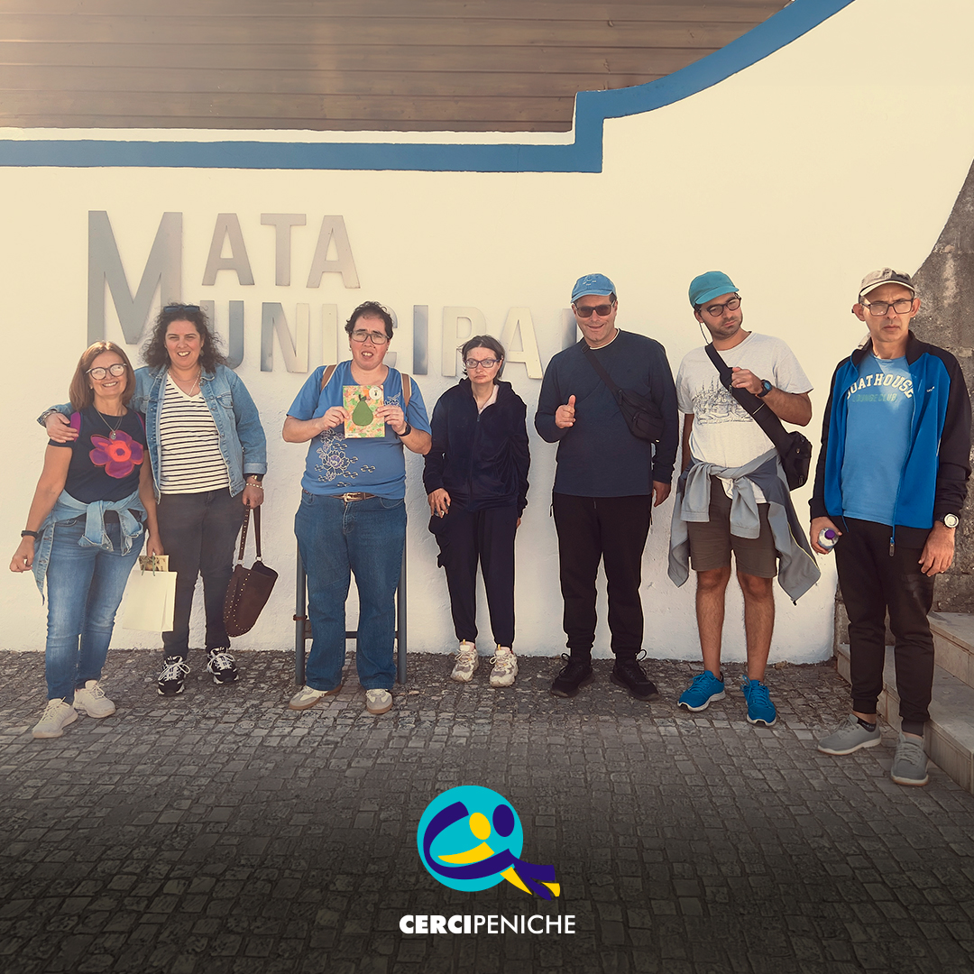6 pessoas apoiadas e monitor da Cercipeniche posam para uma fotografia de grupo junto à entrada da Mata Municipal do Bombarral. Ao fundo, vê-se uma parede branca com a inscrição «Mata Municipal». Na parte inferior da imagem encontra-se o logótipo da Cercipeniche.